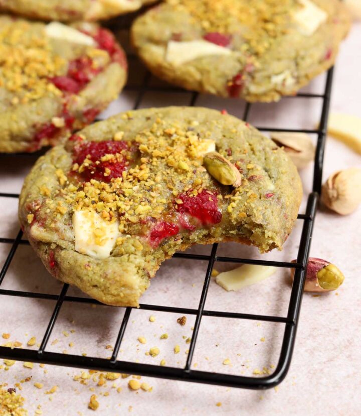 angle view of white chocolate raspberry pistachio cookie with bite on cooling rack showing fudgy center