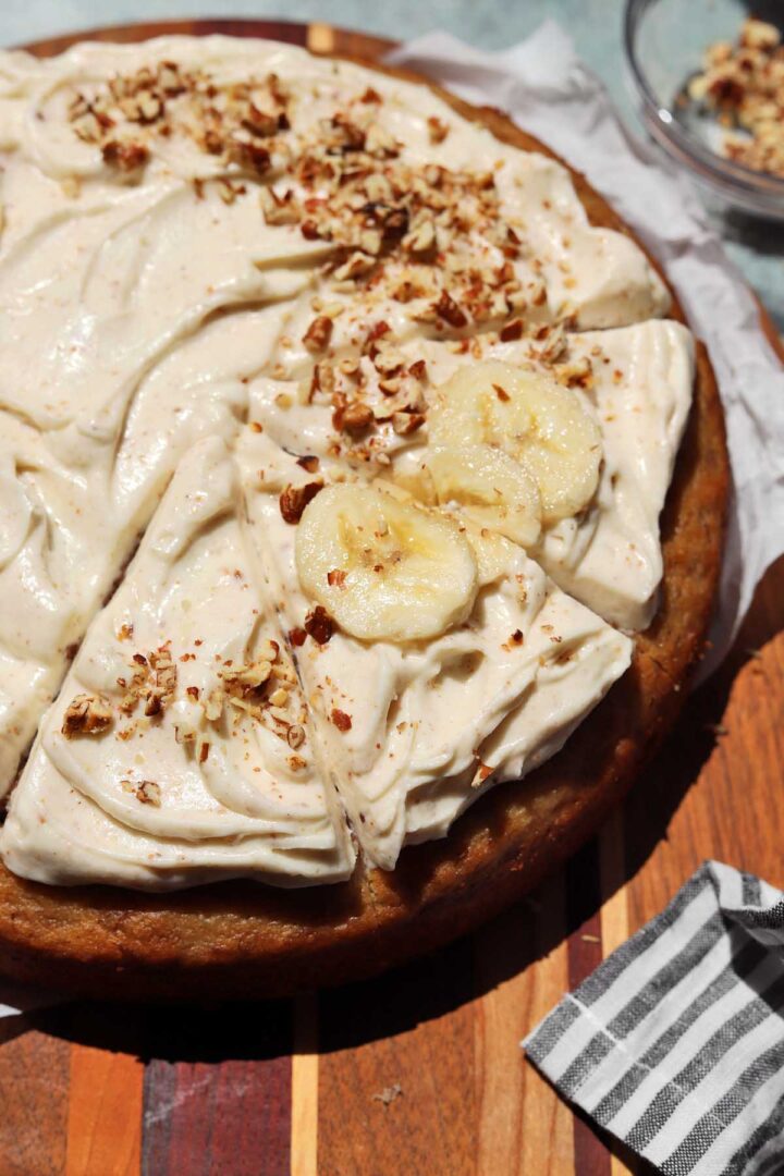 angle view of brown butter banana cake with brown butter cream cheese frosting, sliced bananas and pecans