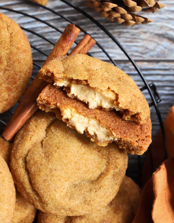 pumpkin cheesecake cookies piled up on cooling rack with cookie split in half showing creamy cheesecake filling