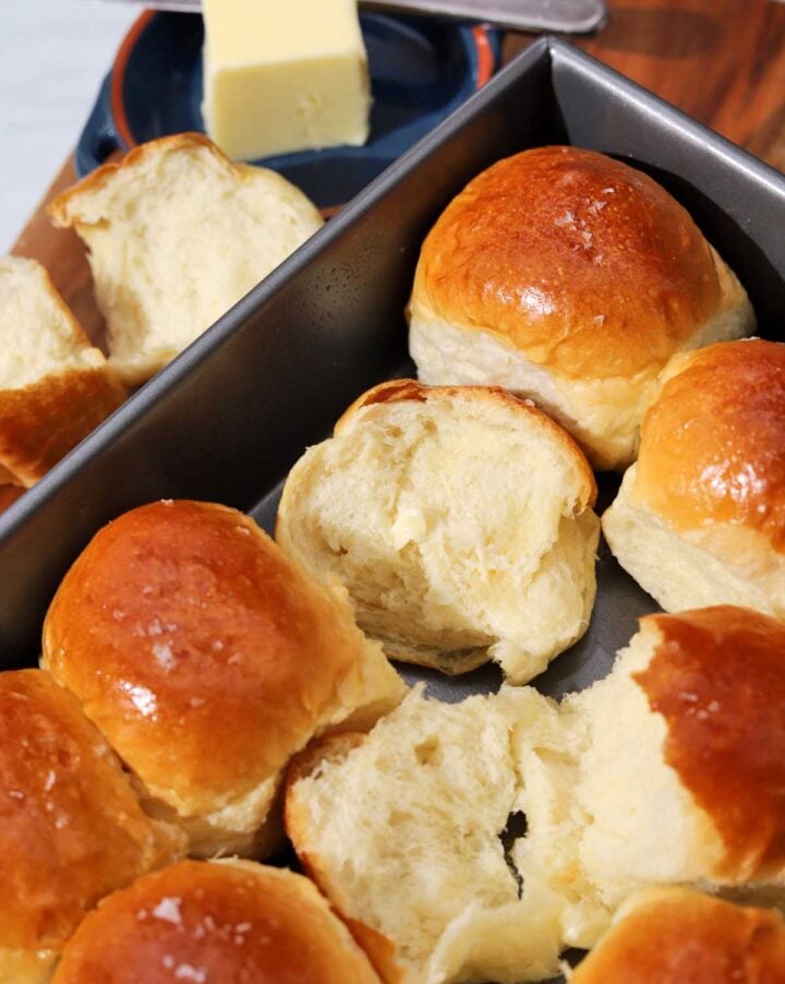 soft homemade dinner rolls in baking pan with one roll torn open with a pad of butter on top