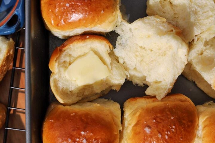 soft homemade dinner rolls in baking pan with one roll torn open with a pad of butter on top